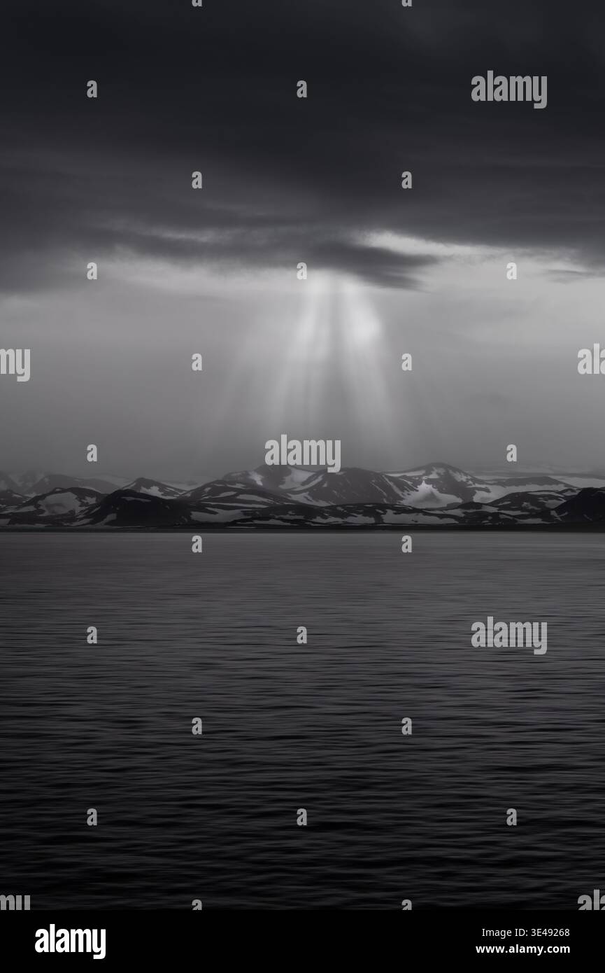 View of dramatic sunbeams breaking through dark storm clouds over the calm sea and snow-covered mountain peaks Greenland. Stock Photo