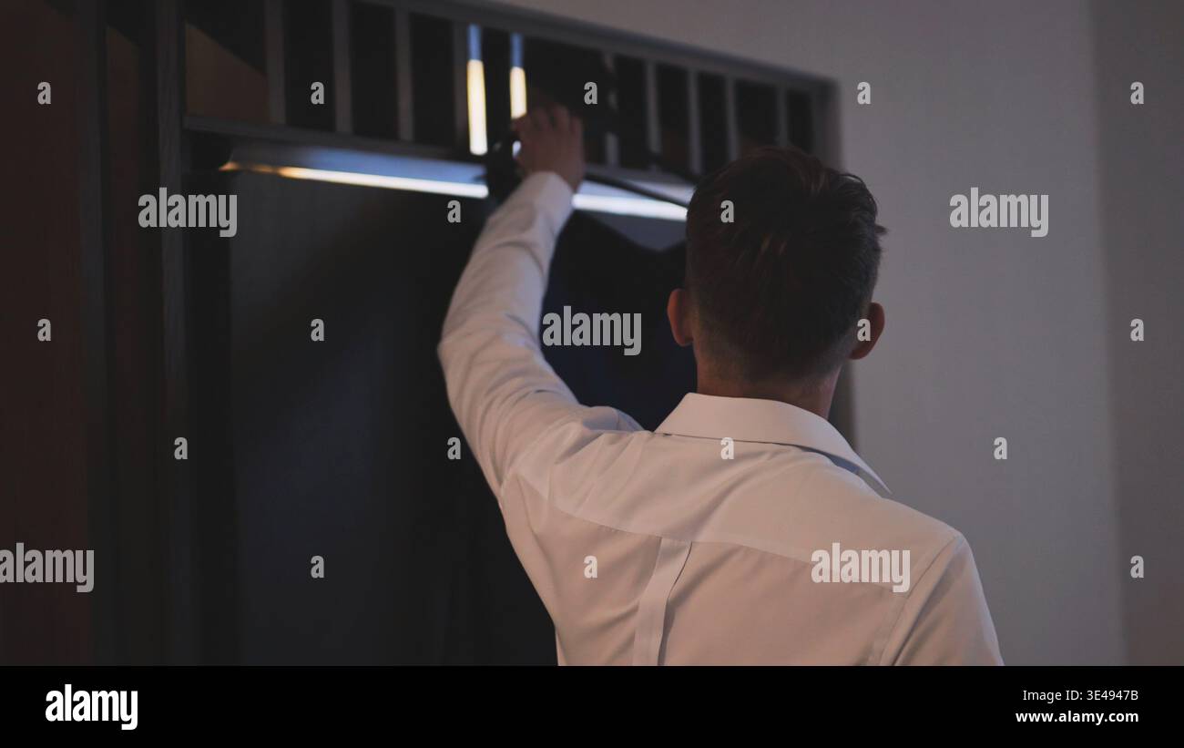 Rear view of a groom in a white dress shirt getting his suit jacket from a dark wardrobe. Stock Photo