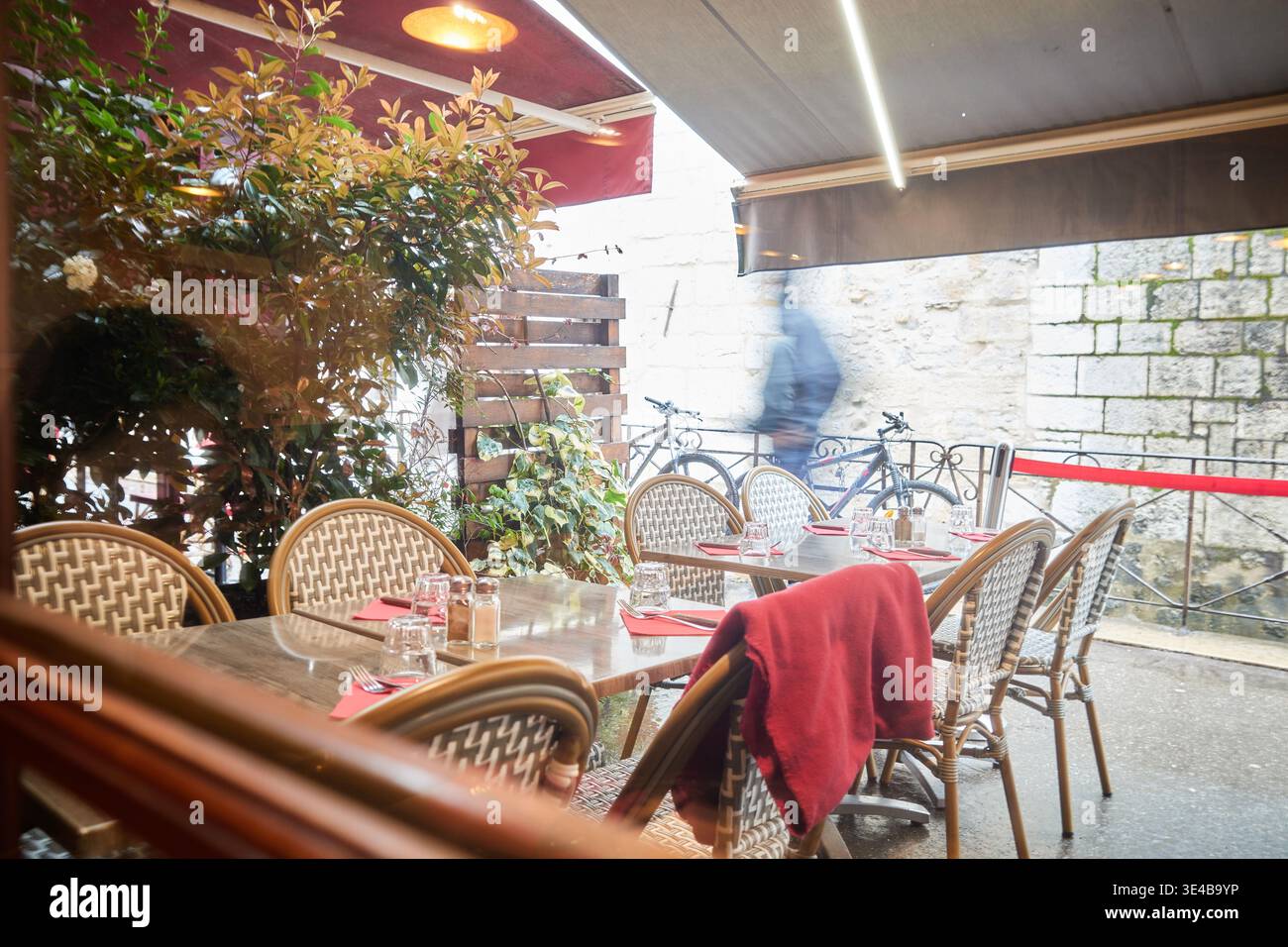 A cozy restaurant terrace with woven chairs and red accents offers a quiet view of a blurred passerby and bicycle against an old stone wall. Stock Photo