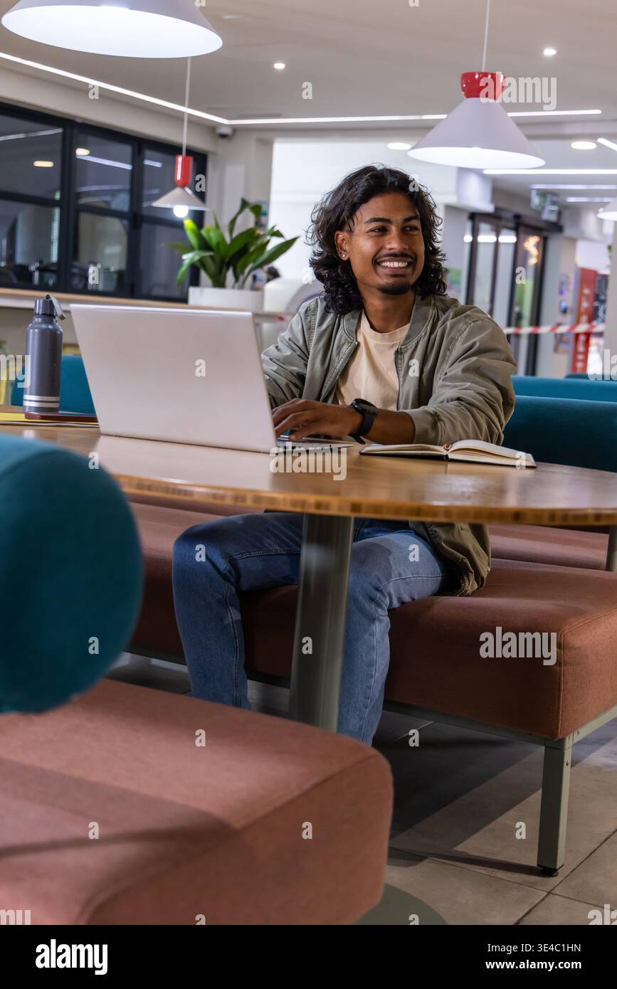 Indian man mid-20s smiling while working on laptop at workspace with notebook, bomber jacket Stock Photo