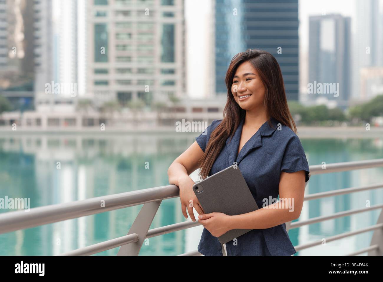 A businesswoman stands by the water in Dubai, smiling confidently while holding a tablet. She represents success in finance. Stock Photo