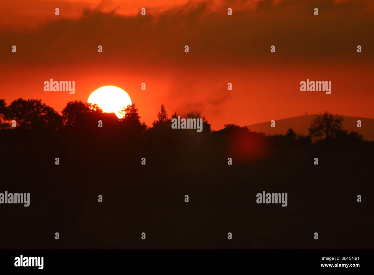 Intense red-orange sunset with the sun partially hidden behind trees and houses. Silhouetted landscape with glowing sky and atmospheric evening light. Stock Photo