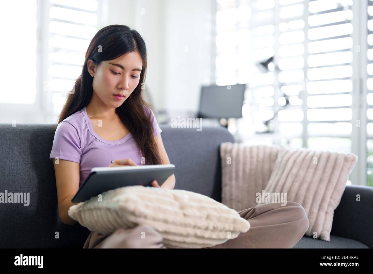 Focused Asian adult woman engaged with tablet at home Modern living room setting shows relaxed remote work or creative study Stock Photo