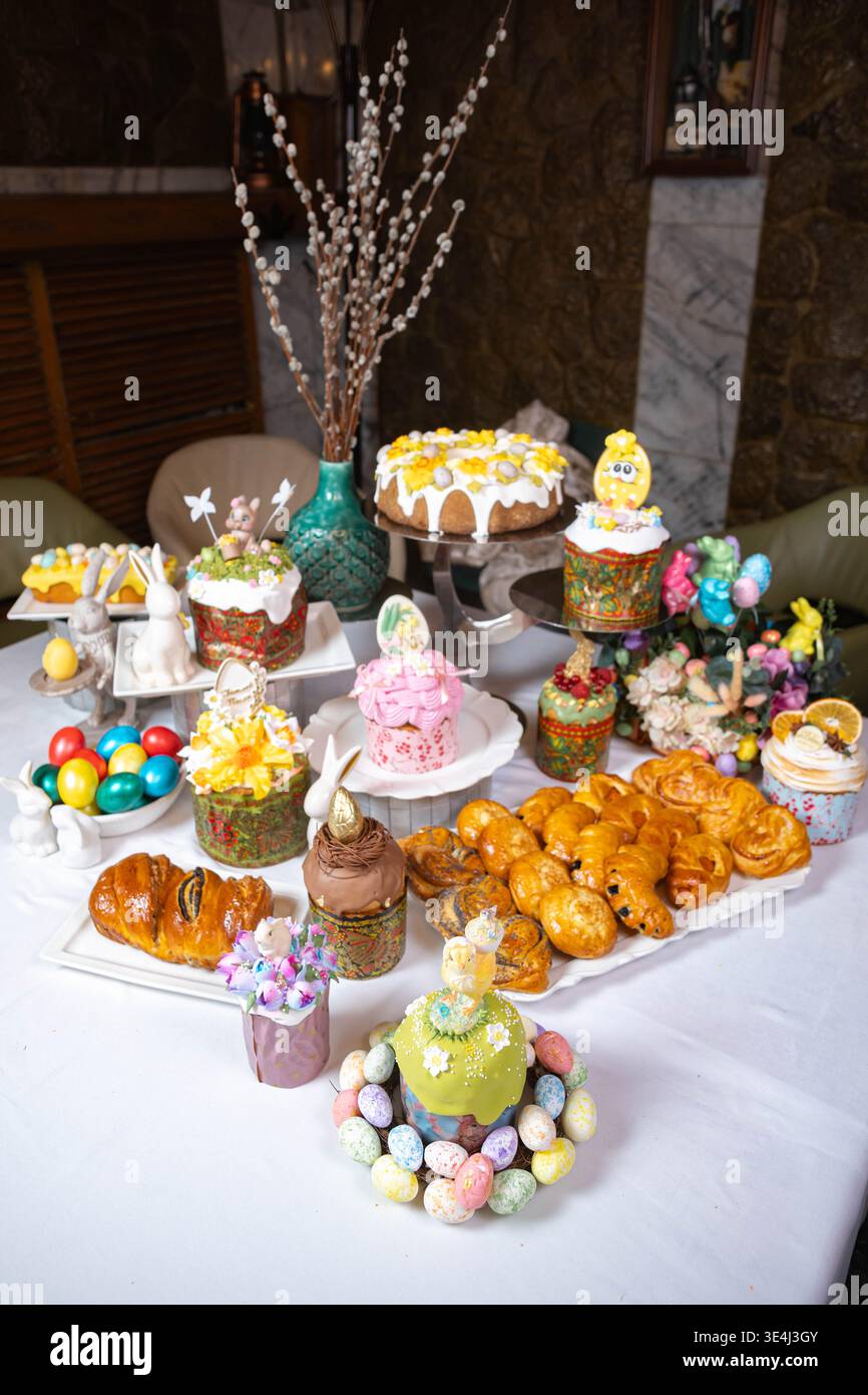 Luxurious Easter table setting on white tablecloth two-tier kulich with floral icing, egg-topped roll, wreath of painted eggs, ceramic bunnies, pastri Stock Photo
