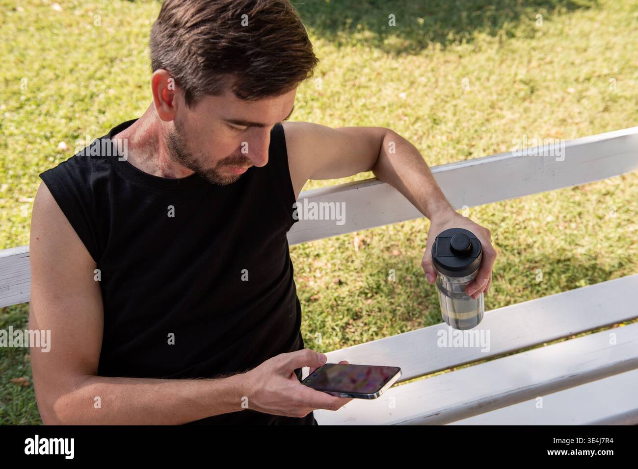 Young man in black sportwear sits on a white park bench after yoga, holding a shaker bottle and checking his smartphone. A red birthmark is visible on Stock Photo
