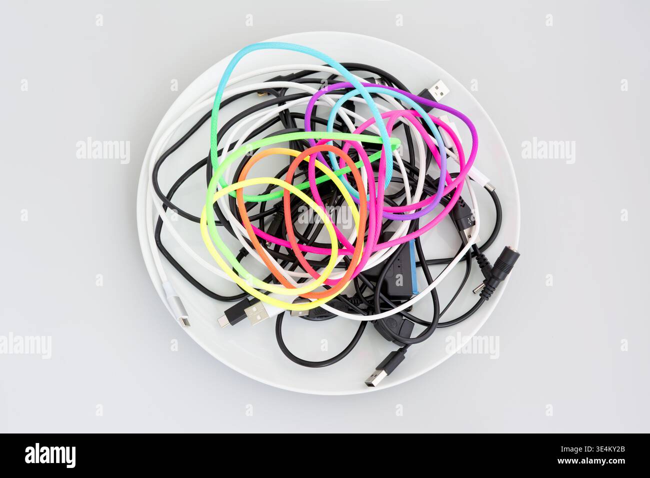 Top view of a white bowl filled with tangled black, white, and brightly colored cables and connectors on a light gray background. Stock Photo
