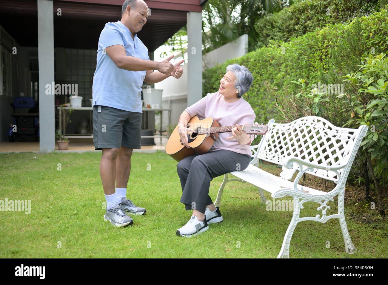 Senior woman sitting on a bench in the garden playing an acoustic guitar while a friend teaching. Concept of music learning and creative leisure in retirement || Model released Stock Photo