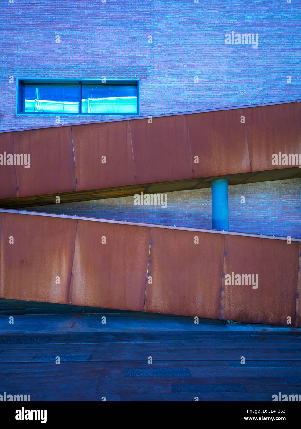 A diagonal rusty corten steel ramp structure crossing a red brick building facade illuminated by vibrant blue lighting at night. The scene represents Stock Photo