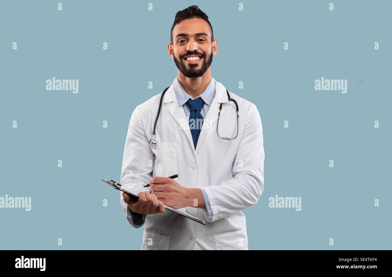 Happy Middle Eastern Doctor Man Taking Notes Standing Over Blue Background Stock Photo