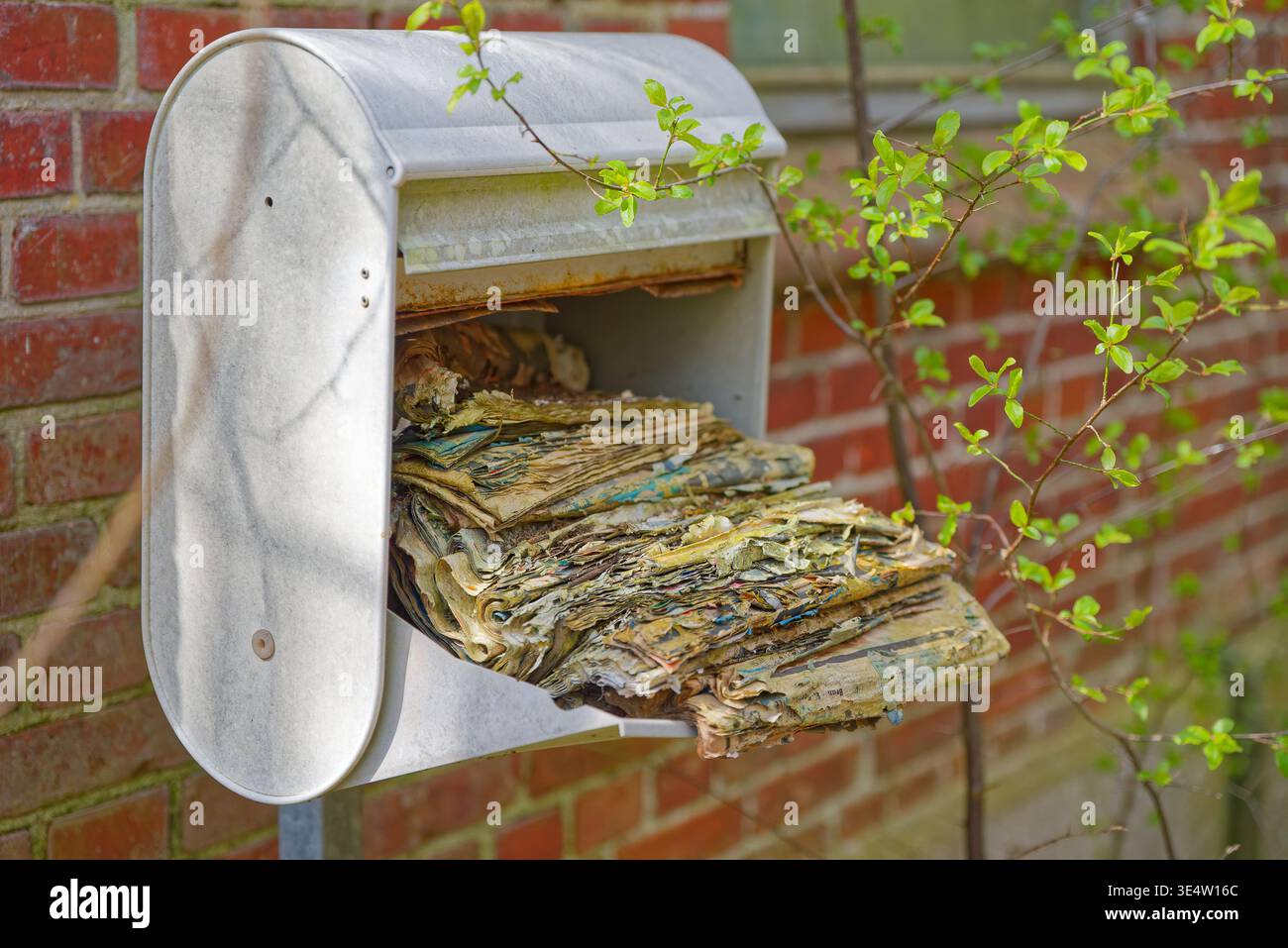 Rusty metal mailbox attached to brick wall filled with old weathered newspapers showing neglect and exposure to elements outdoors. Stock Photo