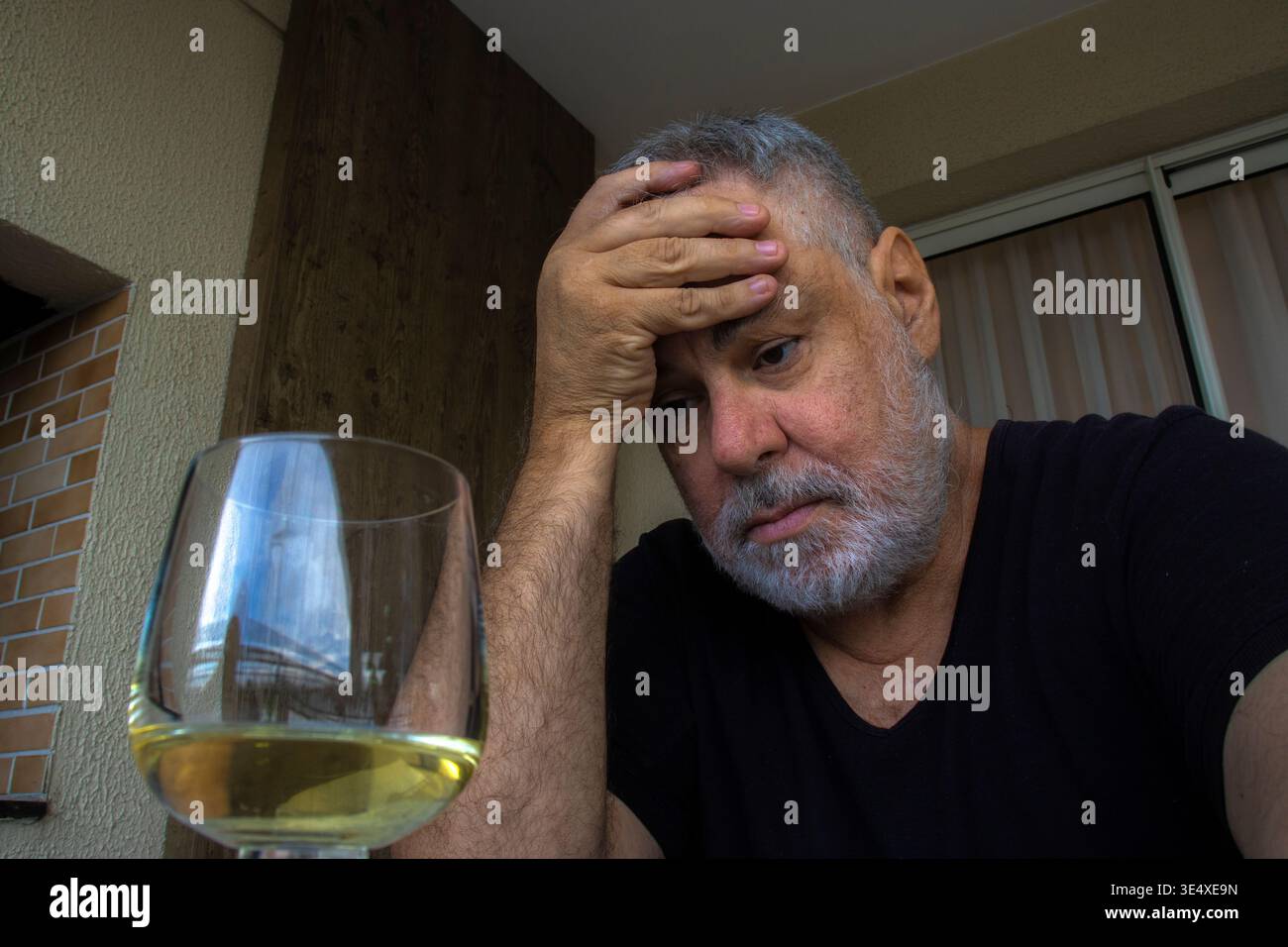 Adult man showing signs of emotional distress rests his hand on his head while looking at a glass containing an alcoholic beverage. The indoor setting Stock Photo