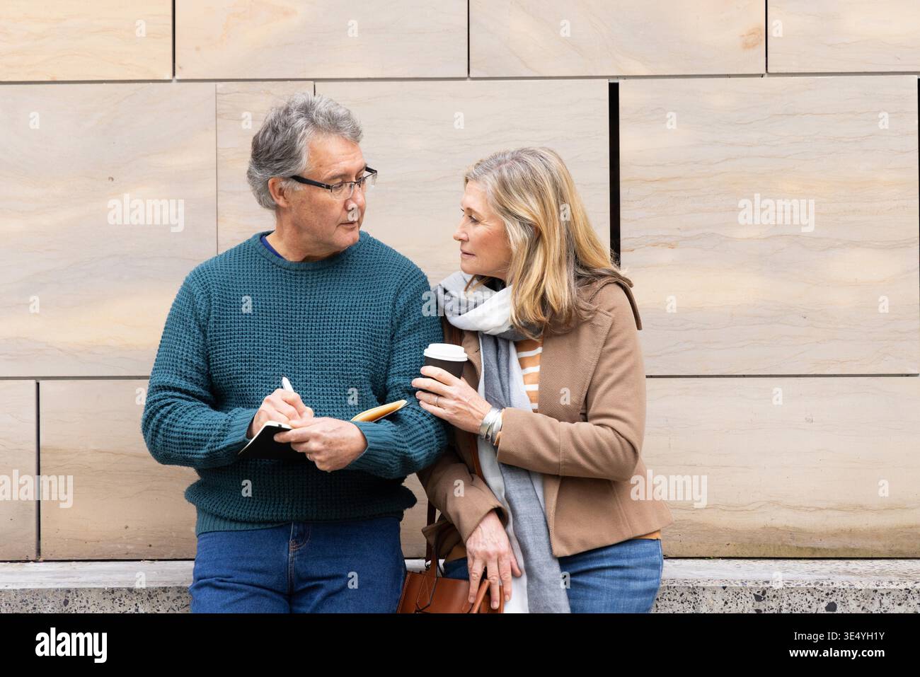 Senior couple standing against beige wall on street, man writing, woman holding coffee. Urban, duo, notebook, pen, scarf, coat, handbag || Model released Stock Photo