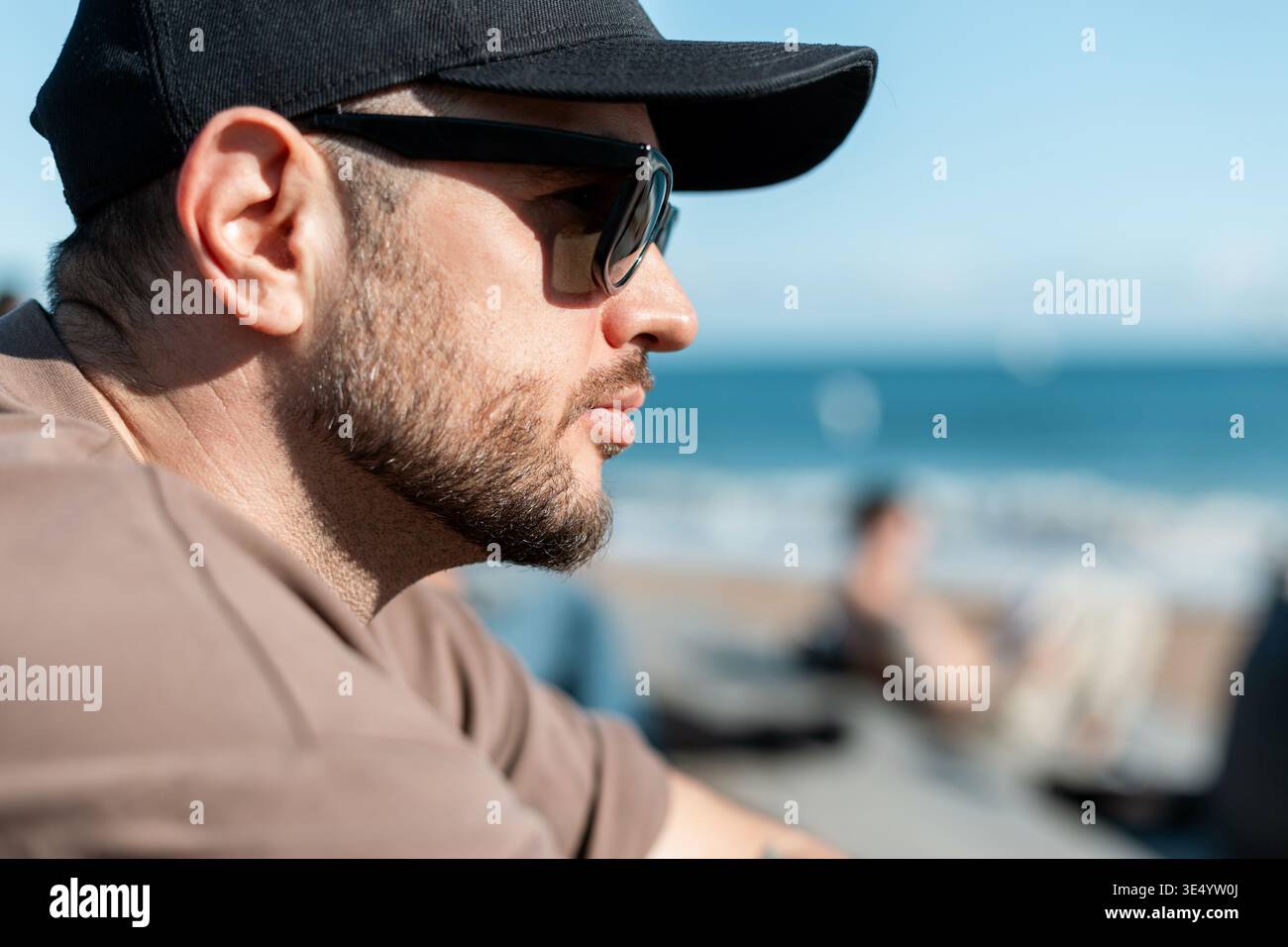 Handsome man with beard with fashion sunglasses with black cap in beige T-shirt sits on beach on seashore. Stock Photo
