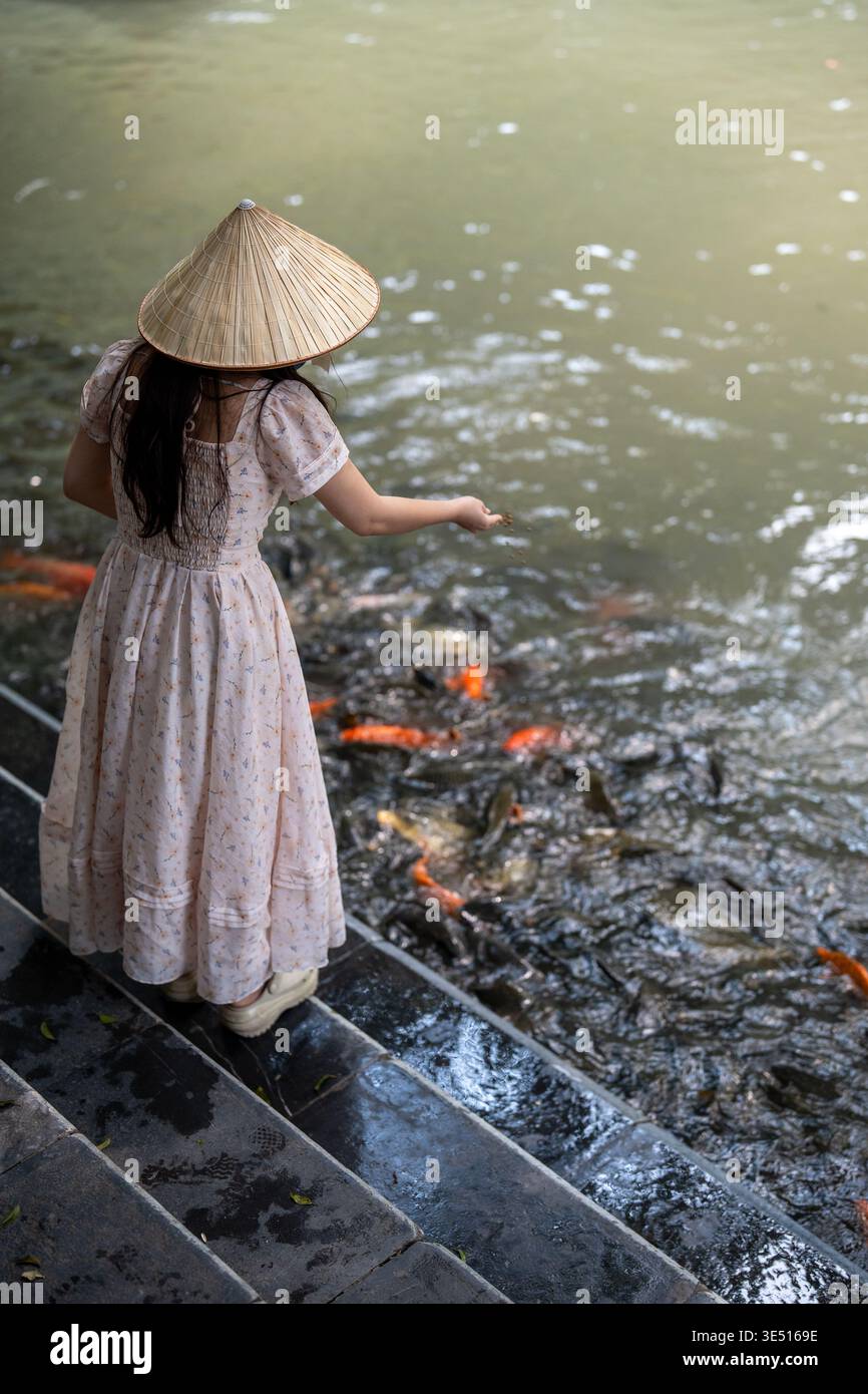 Asian woman in conical hat and fancy dress enjoying feeding freshwater carps fish in pond at park. Stock Photo