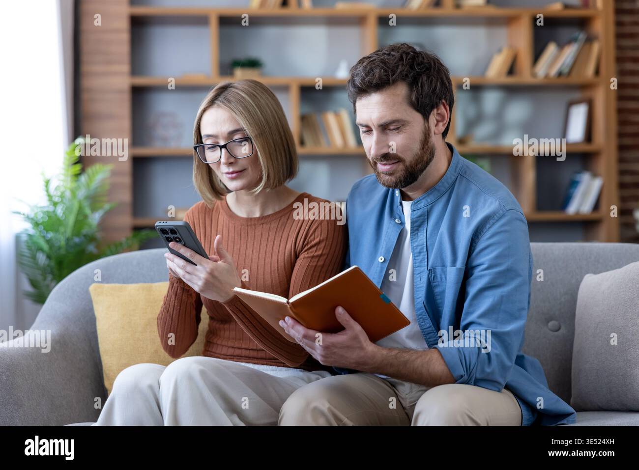 Couple sitting on a couch, collaborating on household tasks, with the woman using a smartphone and the man reading a notebook, both engaged in financial planning and online activities Stock Photo