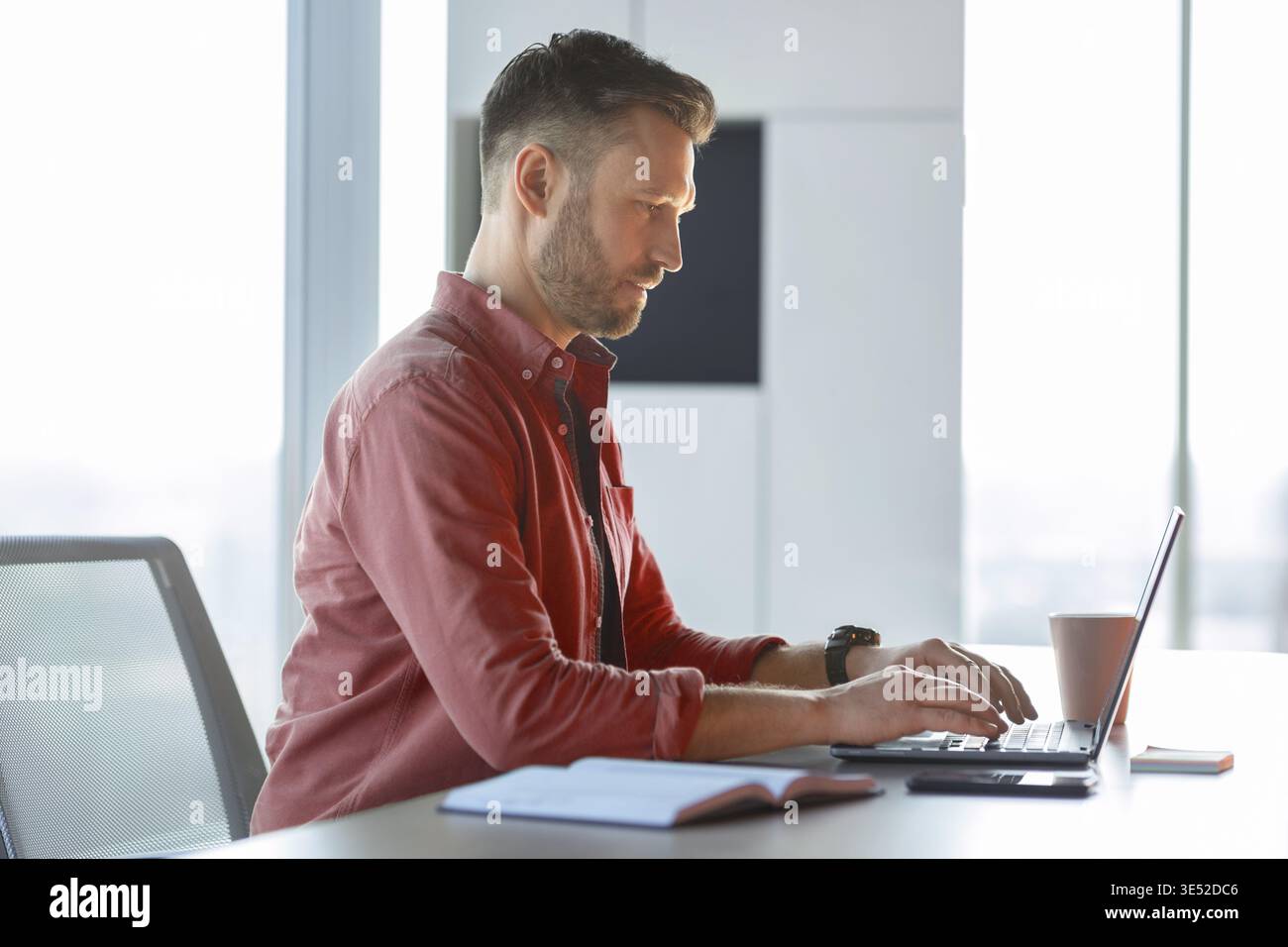 Man Working on Laptop in Bright Office With Cup of Coffee and Notebook on Table Stock Photo