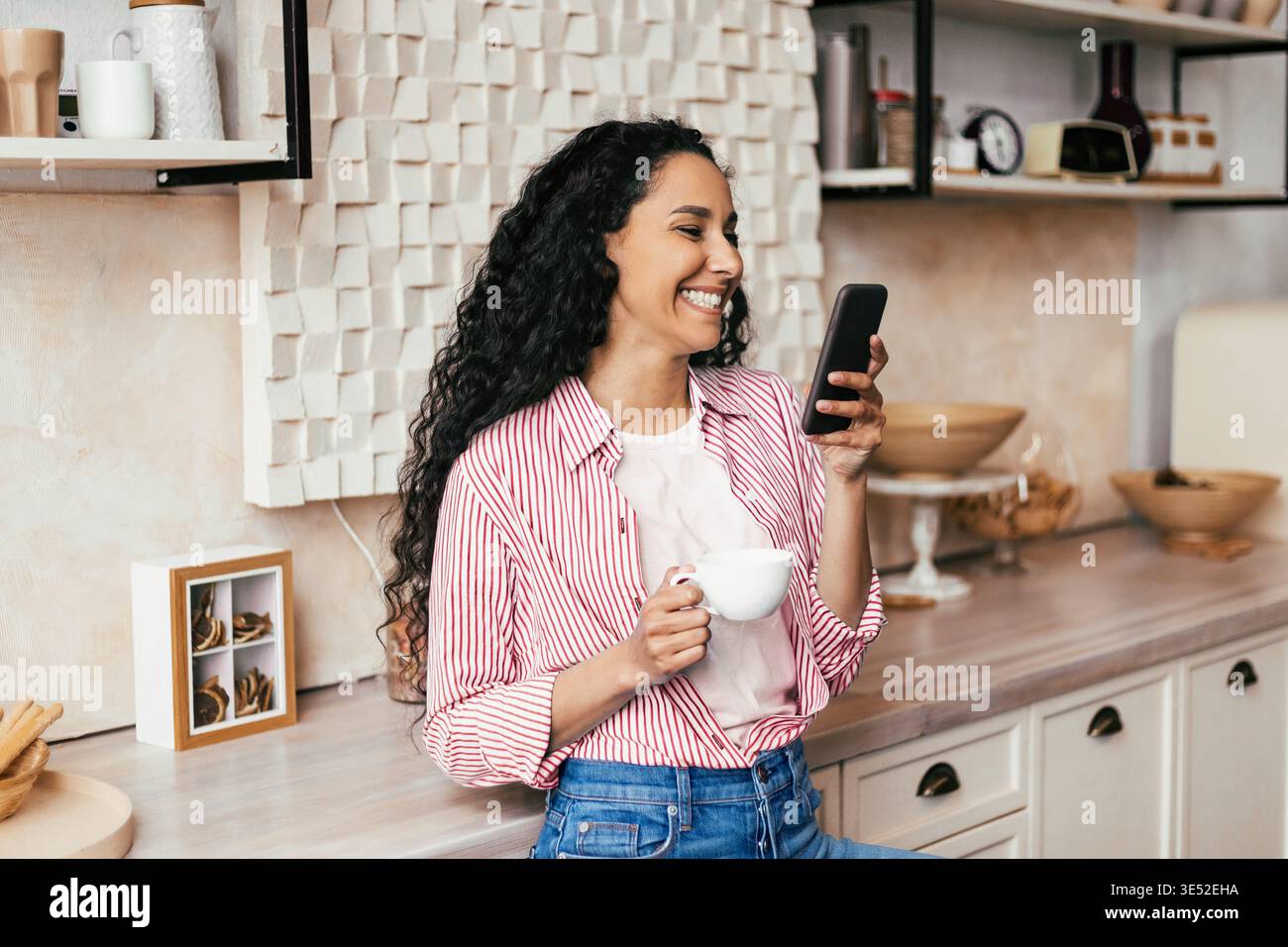 Woman Enjoys Coffee While Using Smartphone in Modern Kitchen Setting Stock Photo