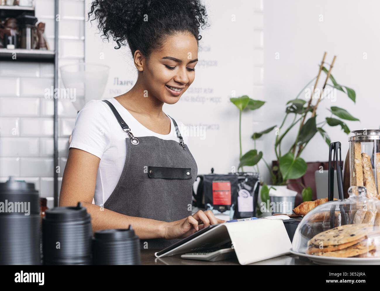 Barista in apron typing on digital tablet. Cheerful woman with curly hair working as a barista in a coffee shop using a digital tablet Stock Photo
