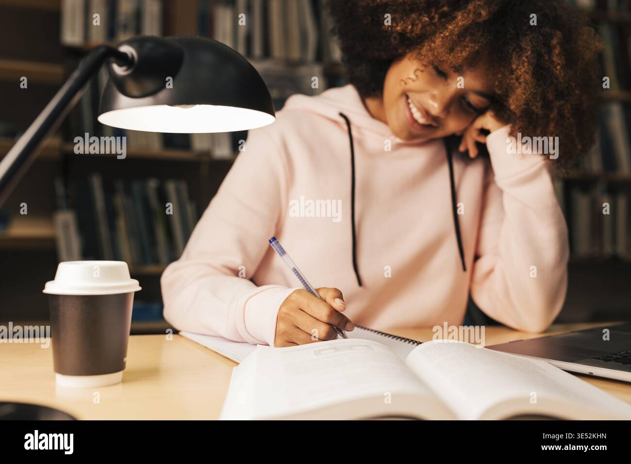 Cheerful student writing on notebook while sitting at desk in library Stock Photo
