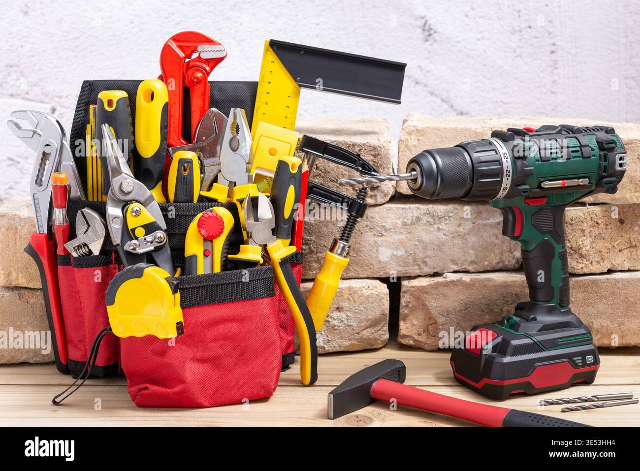 Belt bag with various hand tools and cordless drill with drill bit next to an old brick on a wooden table. Selective focus. Stock Photo