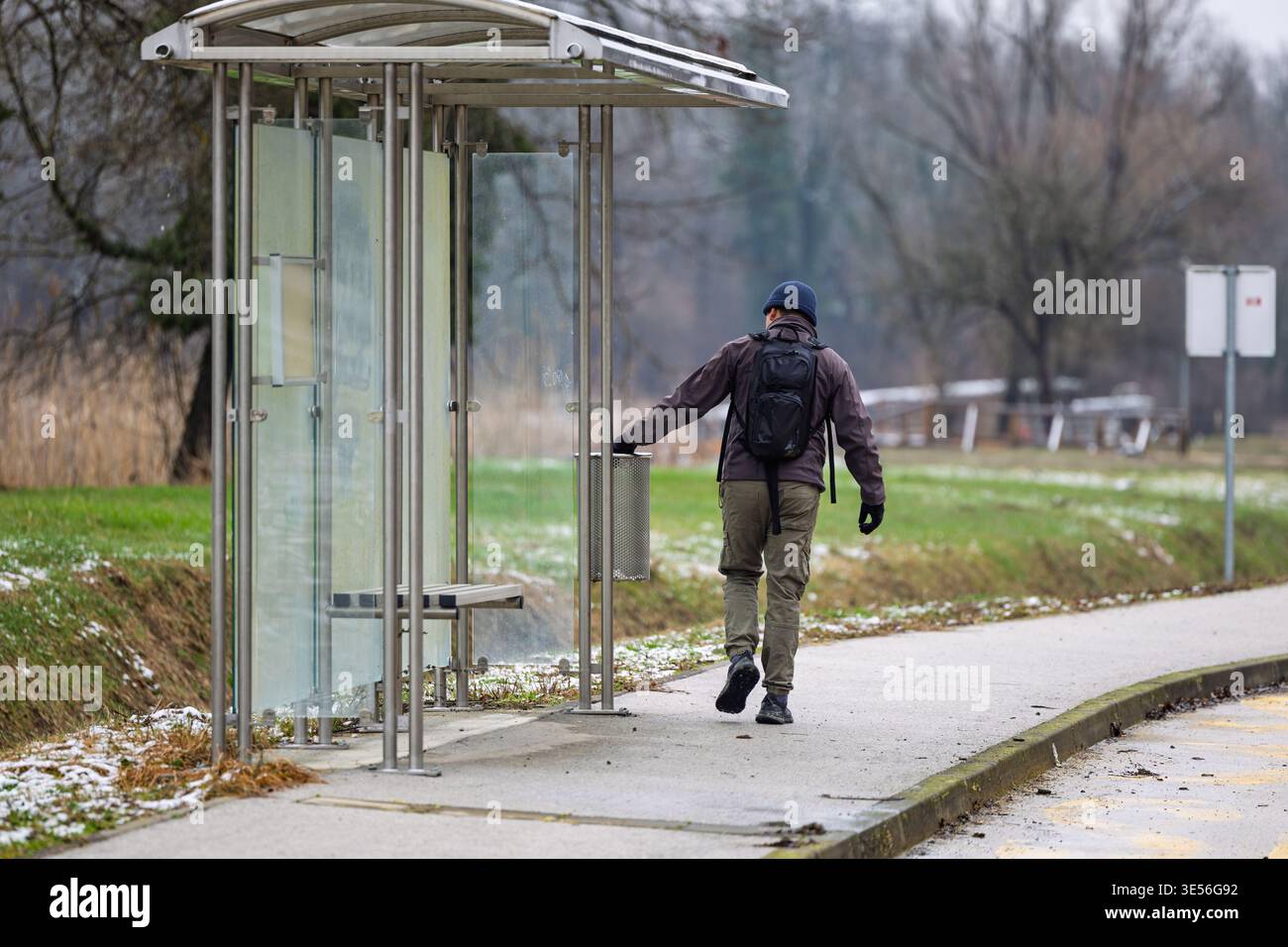 Man wearing a backpack and winter jacket walks past a glass bus stop shelter on an overcast, chilly day, captured from behind along a quiet sidewalk n Stock Photo