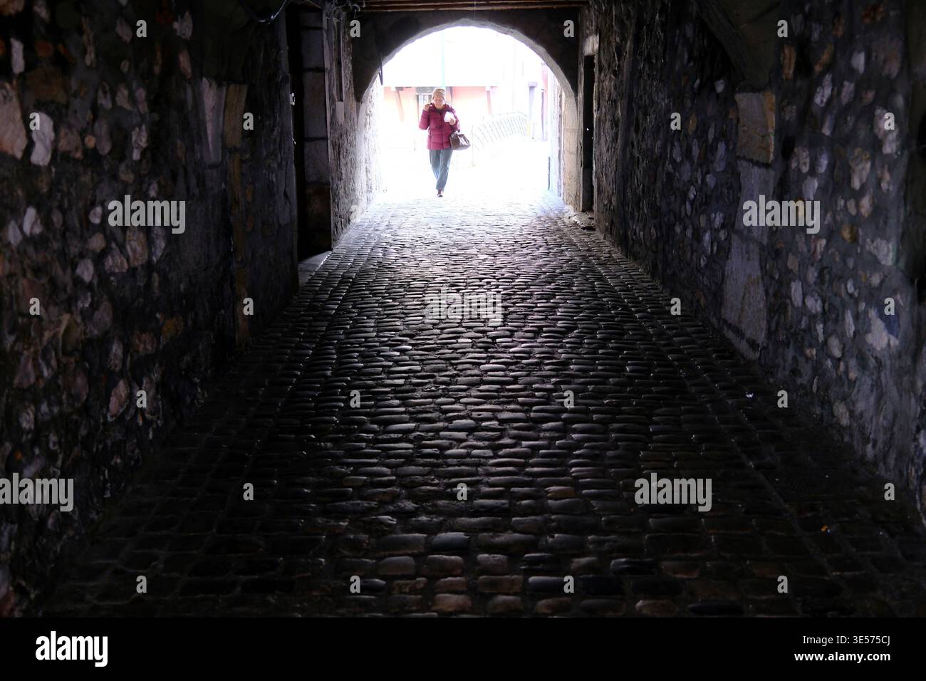 A lone figure emerges from a centuries-old vaulted stone tunnel paved with rounded cobblestones. Annecy. France. Stock Photo