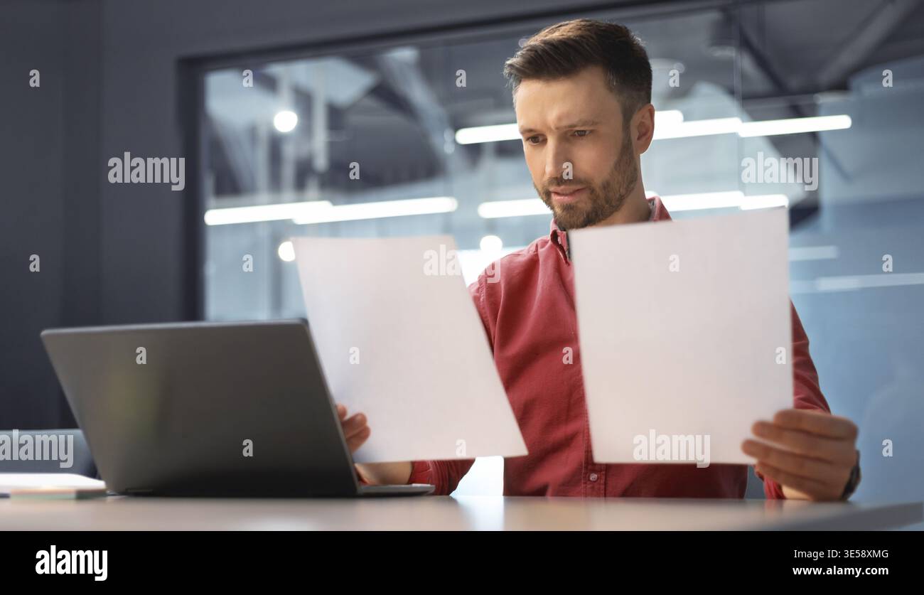 Man Reviews Documents While Using Laptop in Modern Office Setting During Daytime Hours Stock Photo