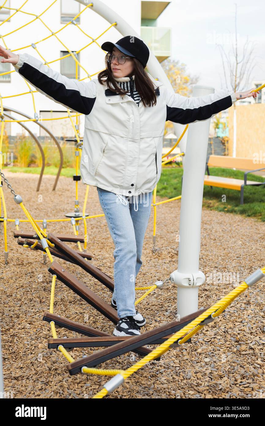 Young woman balancing on a rope bridge in an outdoor playground, expressing playful movement and childlike energy. Stock Photo