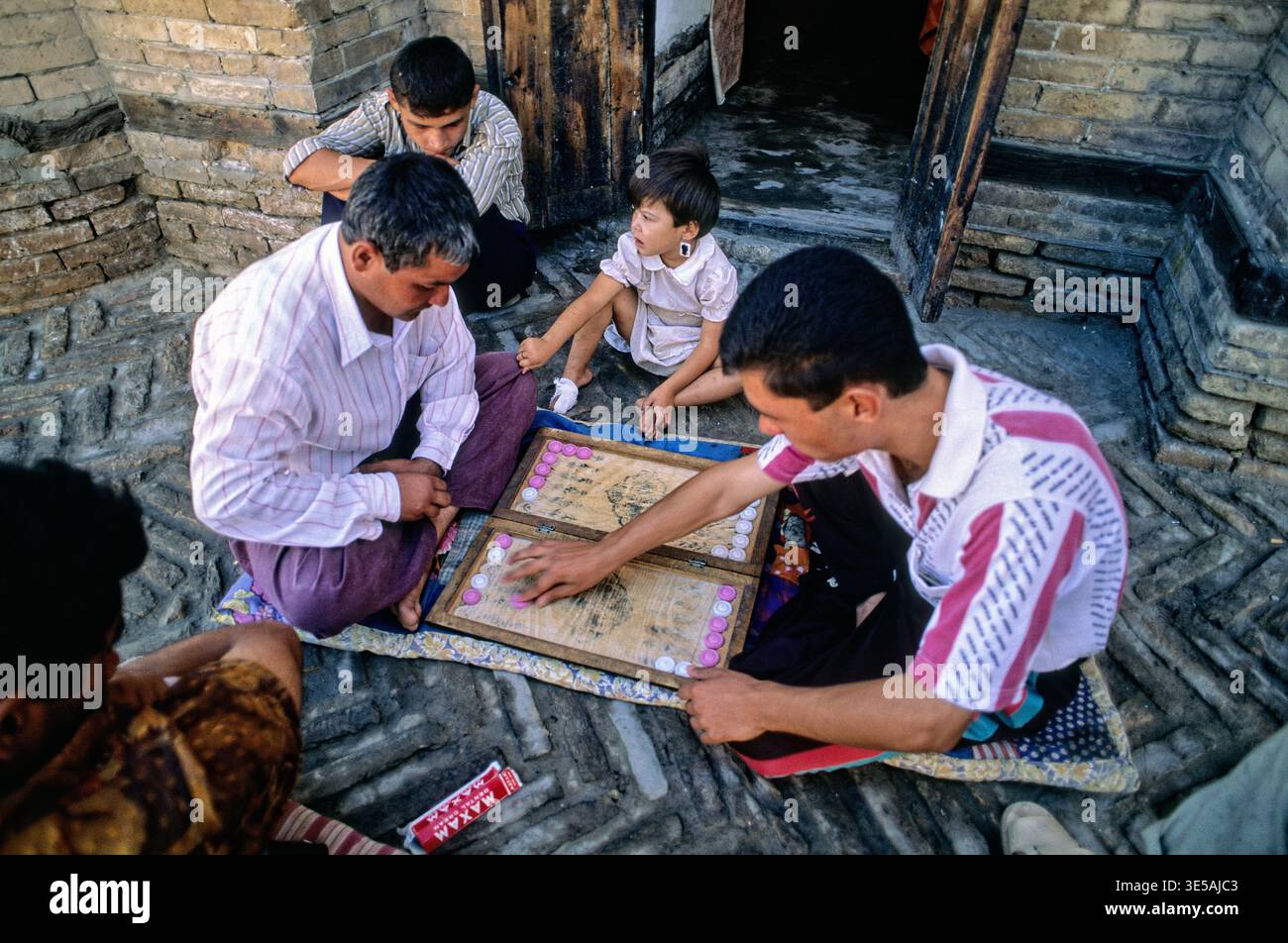 Four men and a child gather on a mat to play a board game in Khiva, Uzbekistan. The setting shows traditional brick walls. Stock Photo