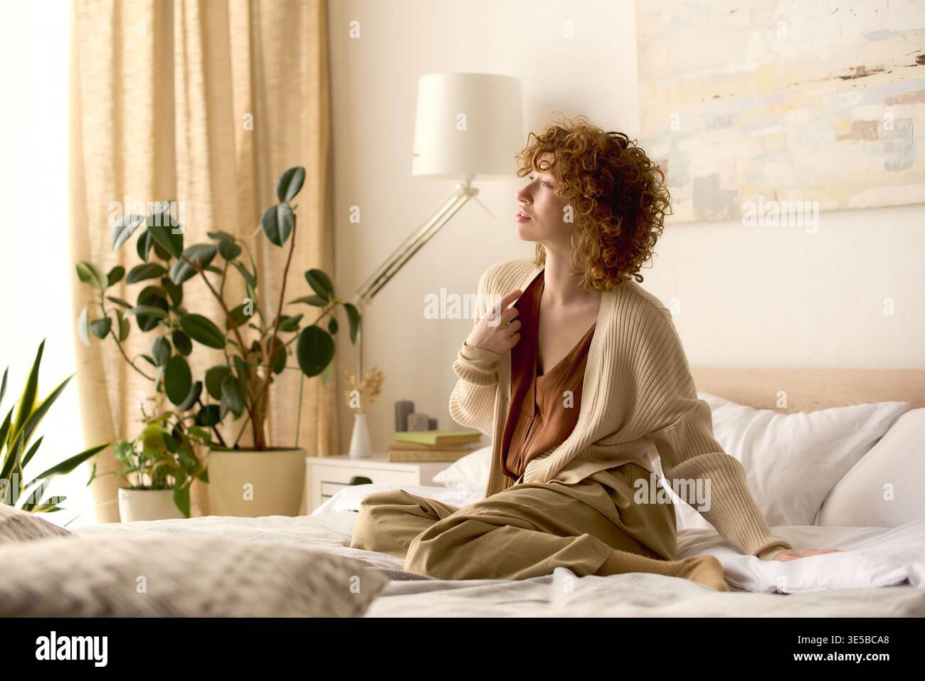 Curly redhead sits peacefully on her bed, surrounded by light and plants. Stock Photo