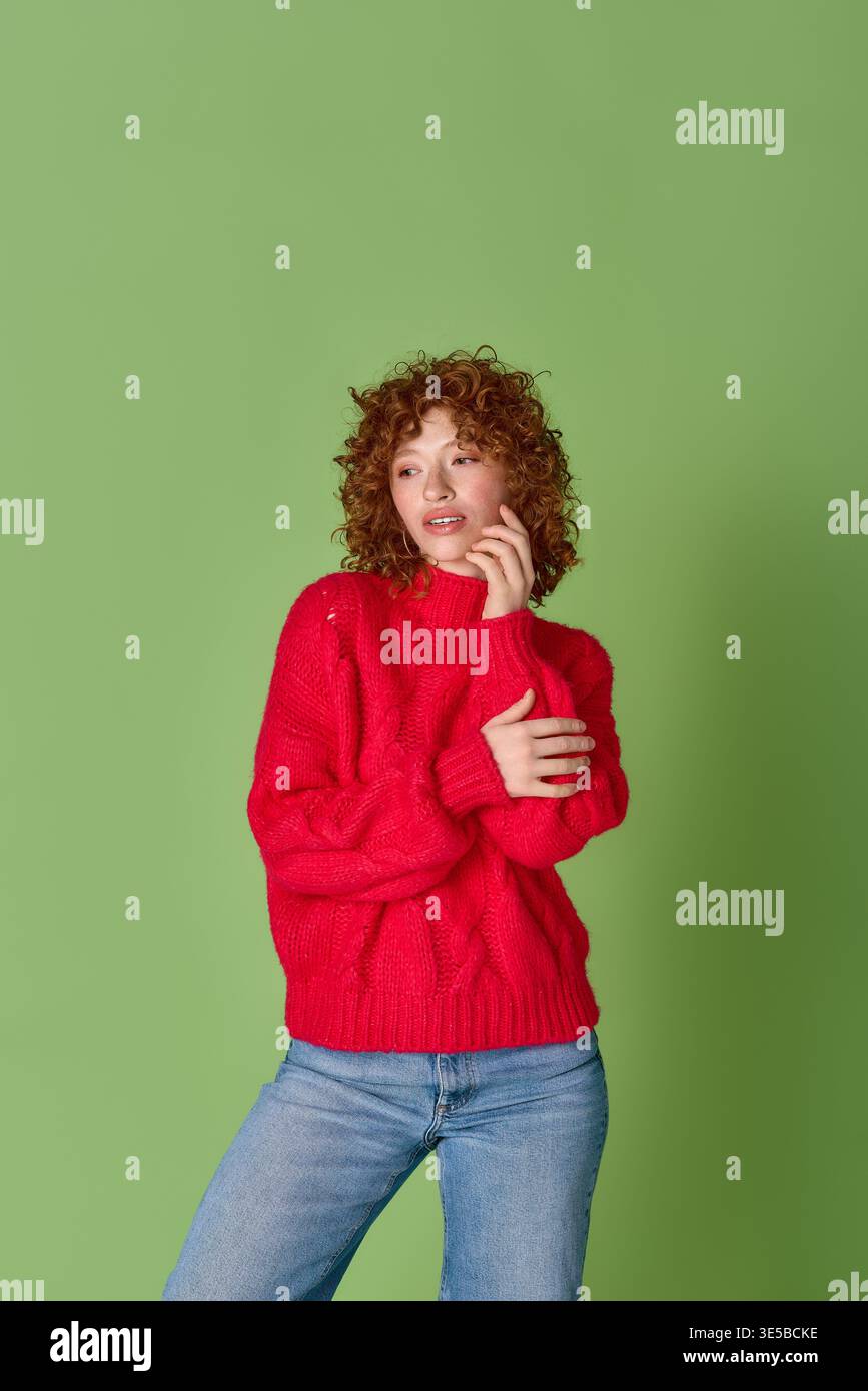 A young woman with stunning red curls poses gracefully in a cozy red sweater on green. Stock Photo