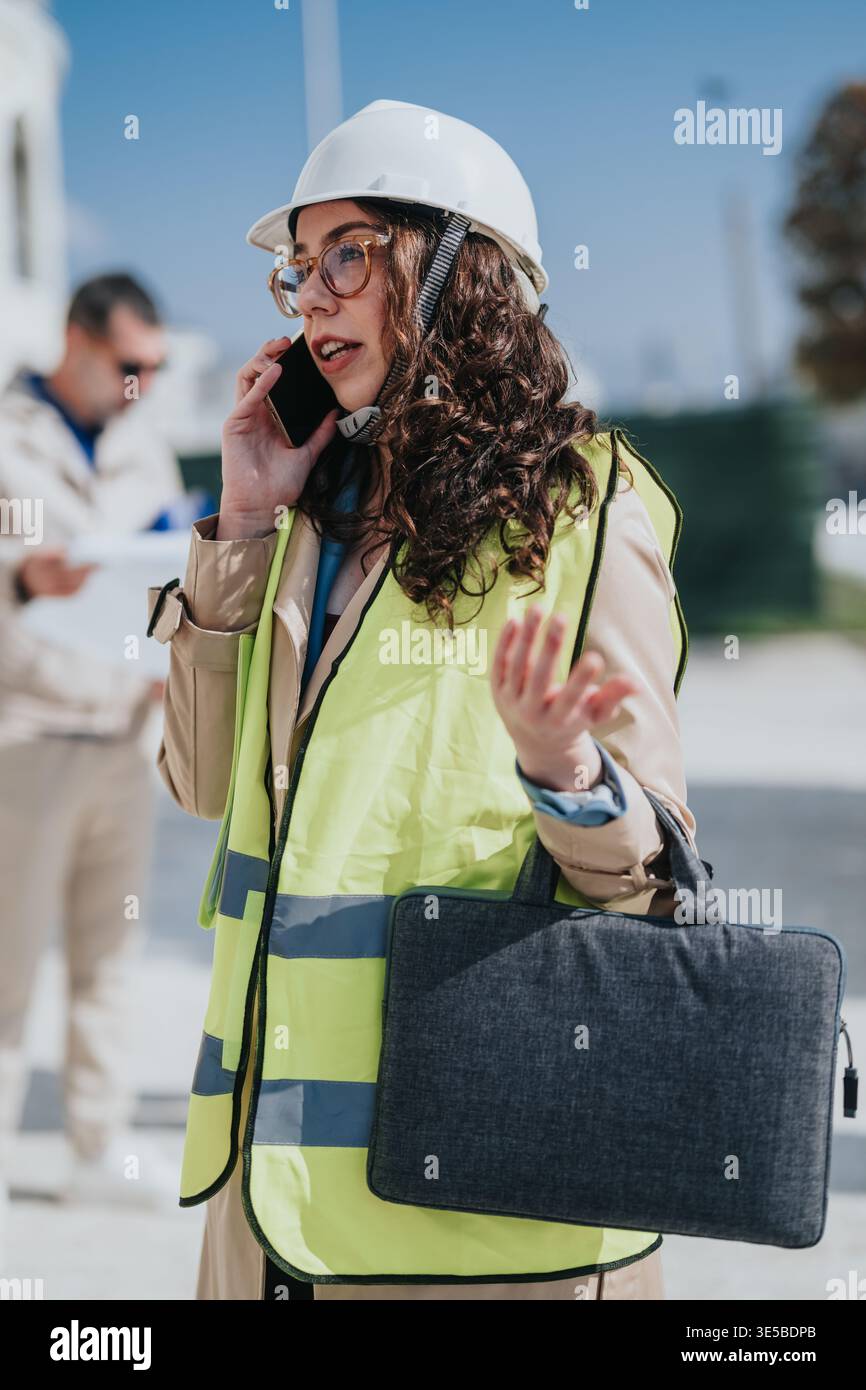 Female construction engineer talking on phone at building site wearing hard hat and safety vest Stock Photo