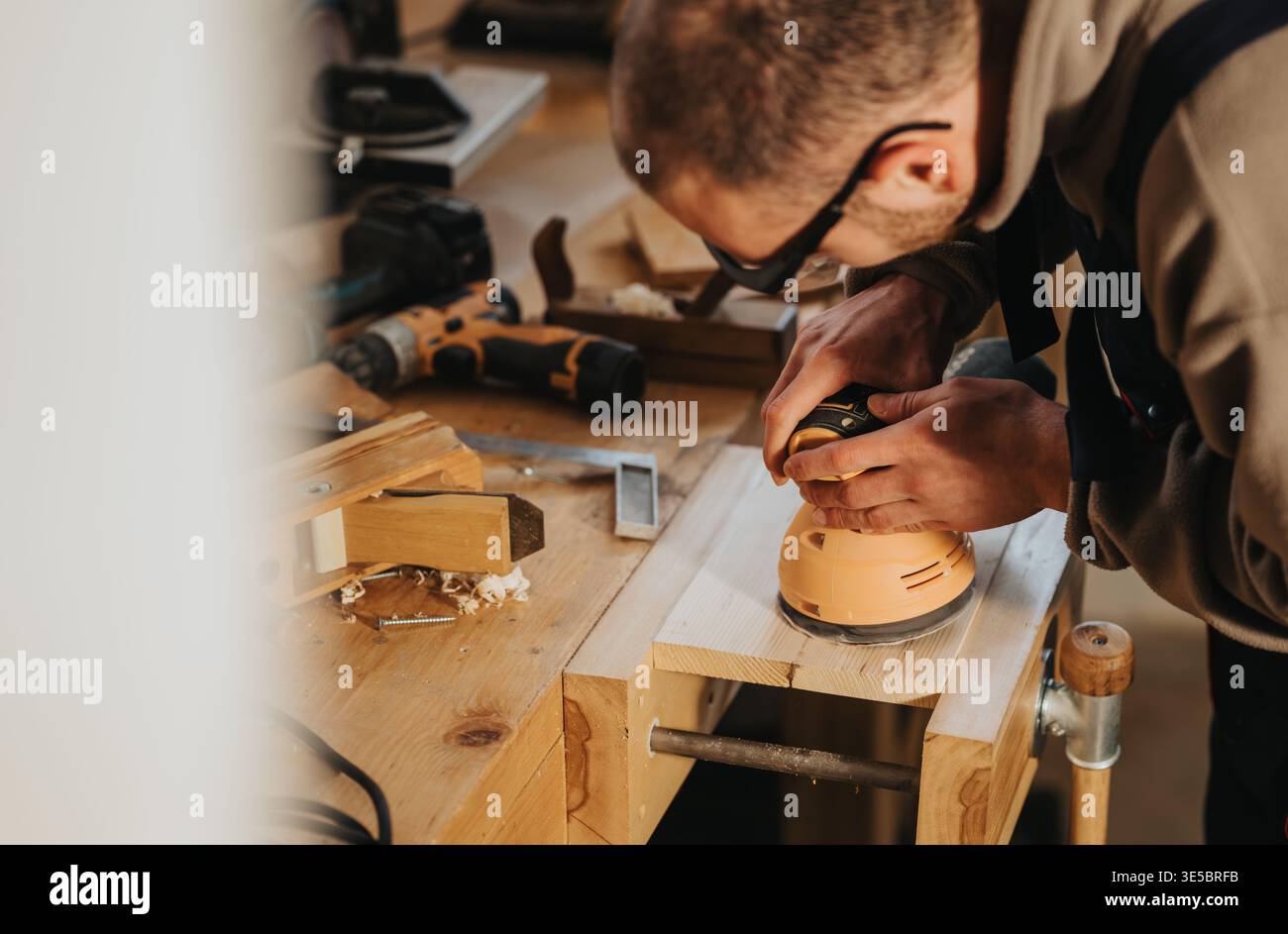 Carpenter sanding wooden plank in workshop using electric sander on workbench Stock Photo