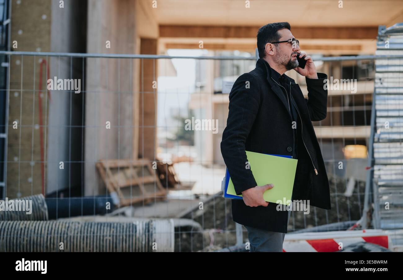 Construction engineer talking on phone while holding project folders at a building site Stock Photo