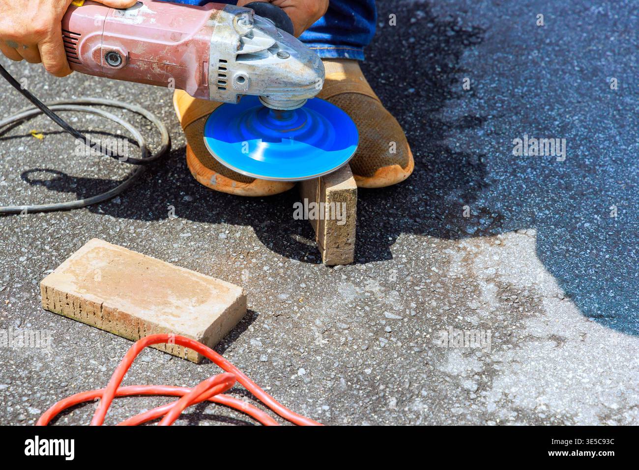 Worker cuts block with grinder tool while standing on dark pavement with other tools, materials around. Stock Photo