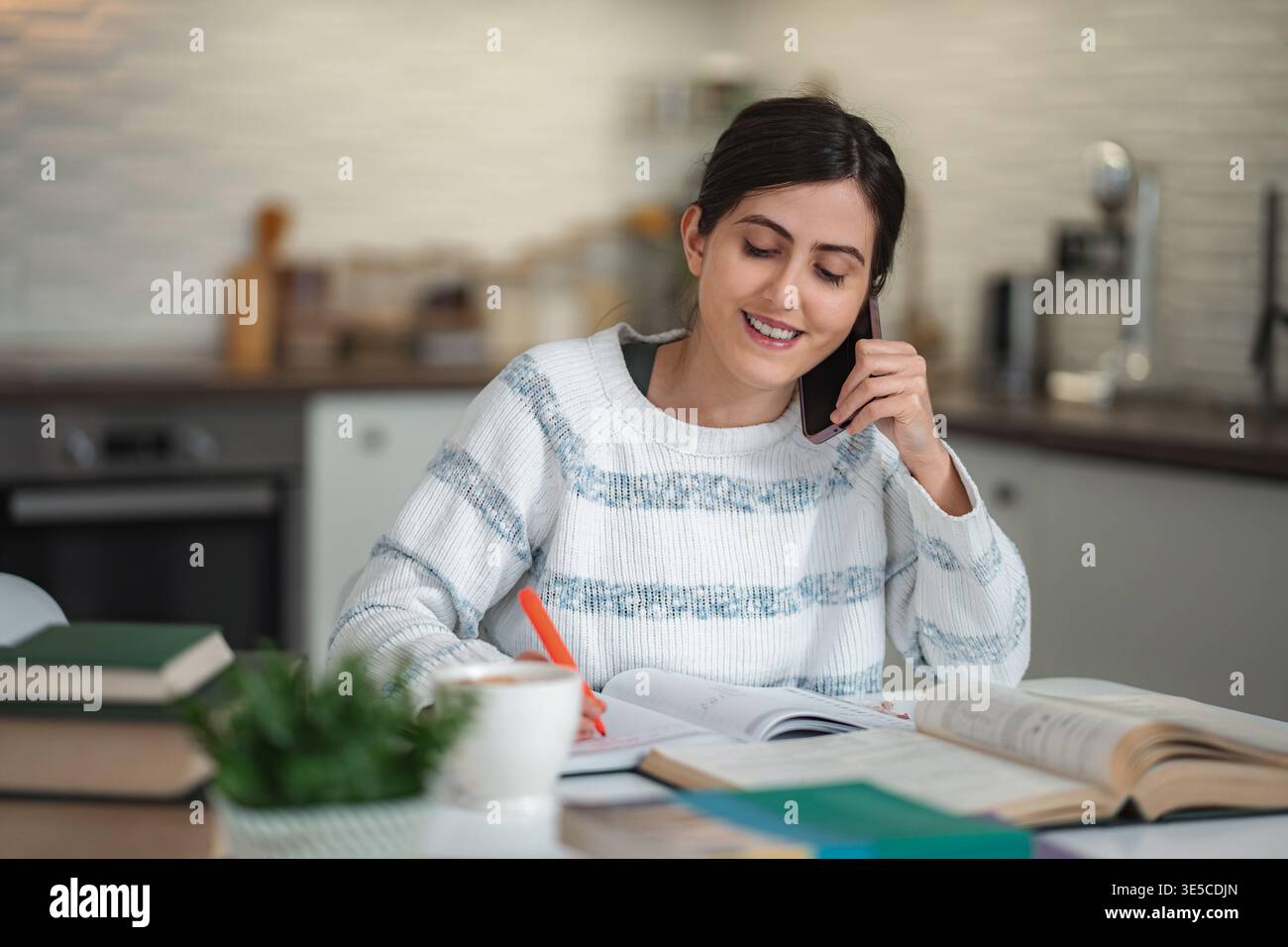 Young woman studying at home while talking on phone Stock Photo
