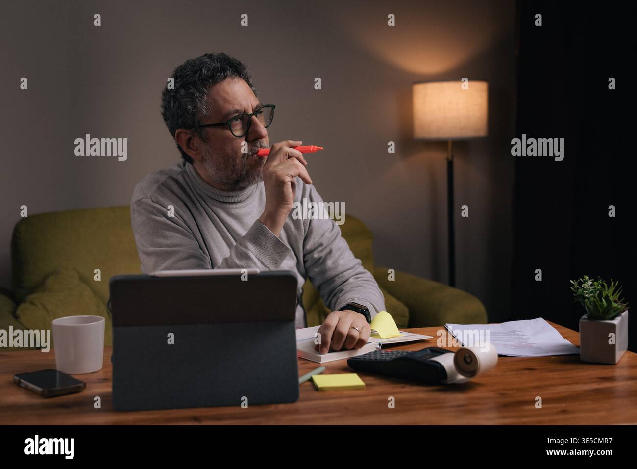 Middle aged man working from home reviewing financial documents and highlighting important information with marker while using tablet. Concept of pers Stock Photo