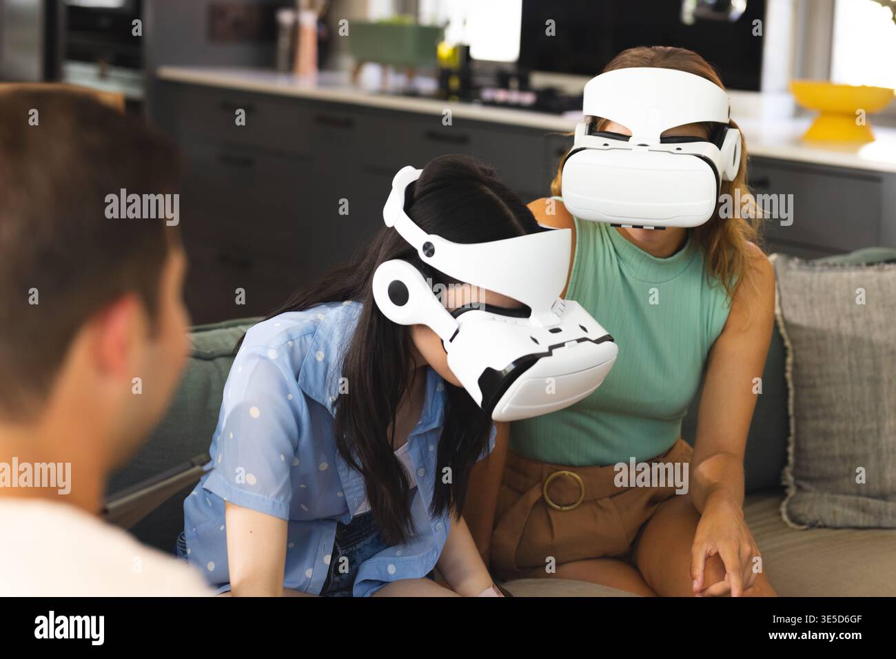 Diverse female friends experiencing VR on grey couch in living room, with white VR headsets Stock Photo