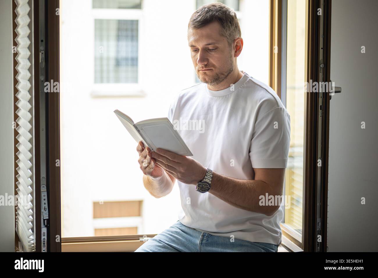 Man reading book while sitting on window sill with natural daylight in modern apartment Stock Photo