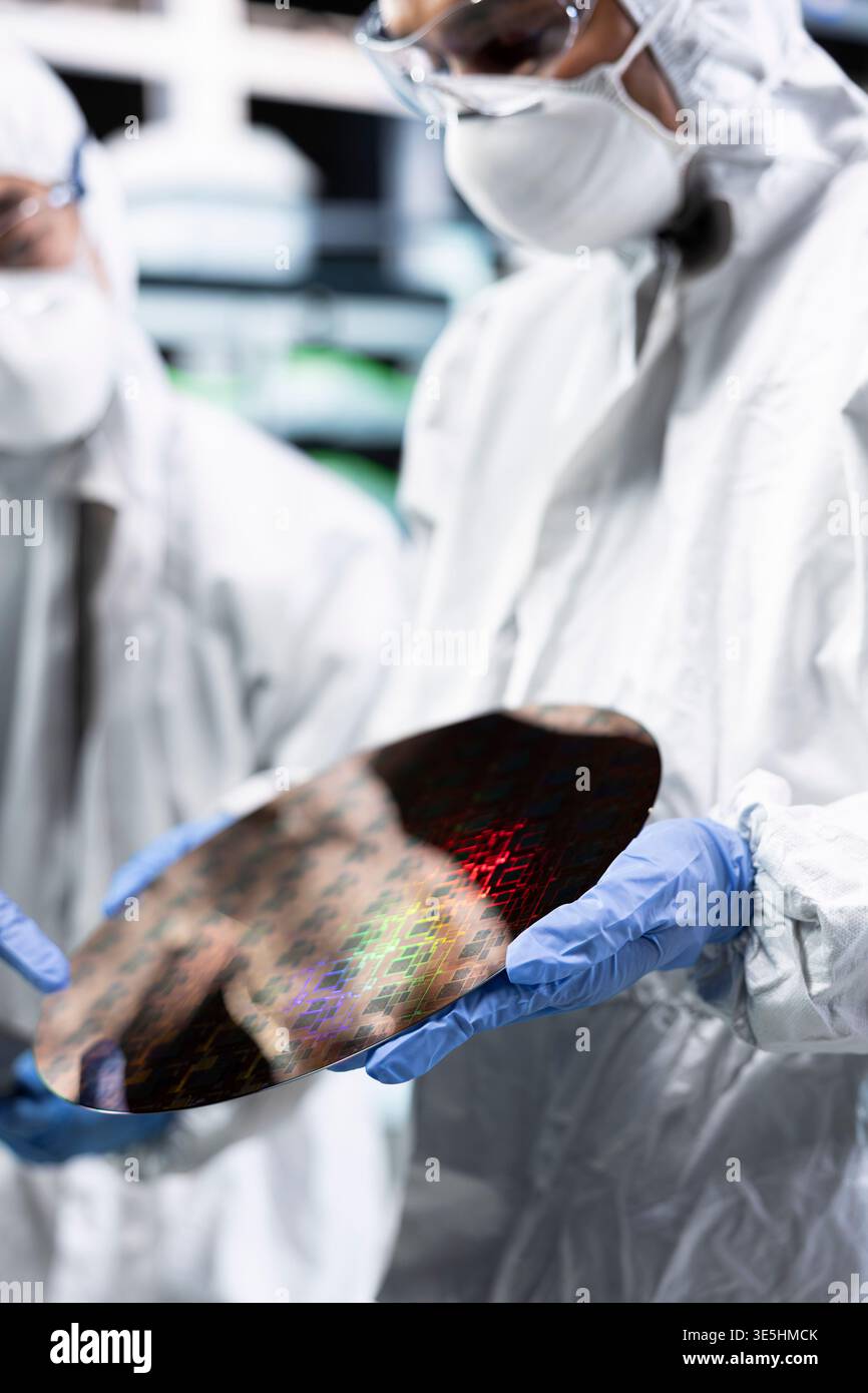 Close up of silicon wafer analyzed by men working together in automated fabrication plant cleanroom. Coworkers in bunny suit examining microelectronic semiconductor substrate for defects Stock Photo