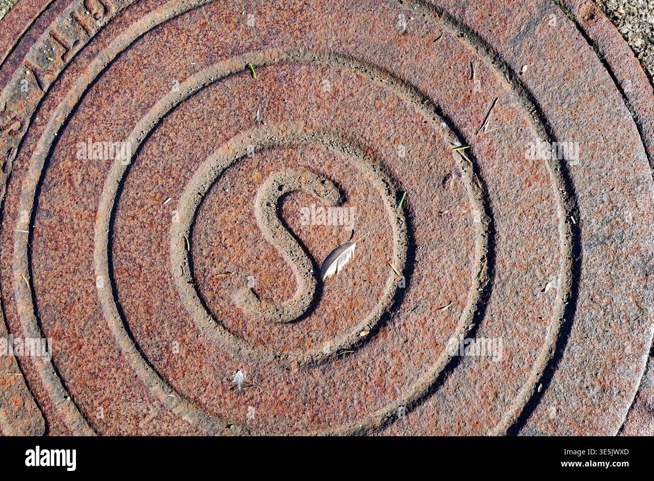 A rusty metal sewer cap on the ground with a small bird feather Stock Photo