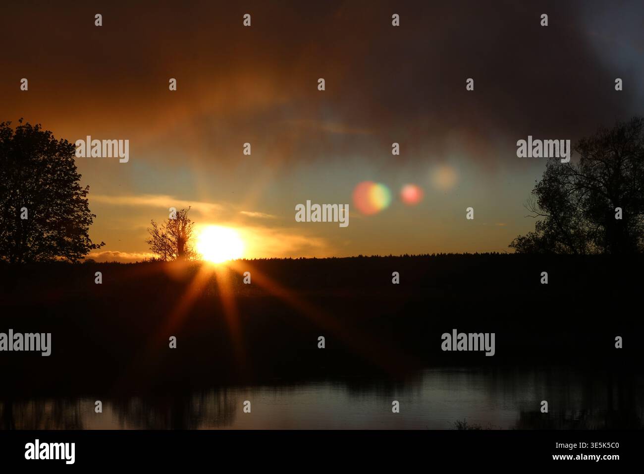 Radiant sunset over a tranquil lake, sunbeams bursting through dark clouds. Silhouetted trees frame the vibrant sky and water reflections with atmosph Stock Photo