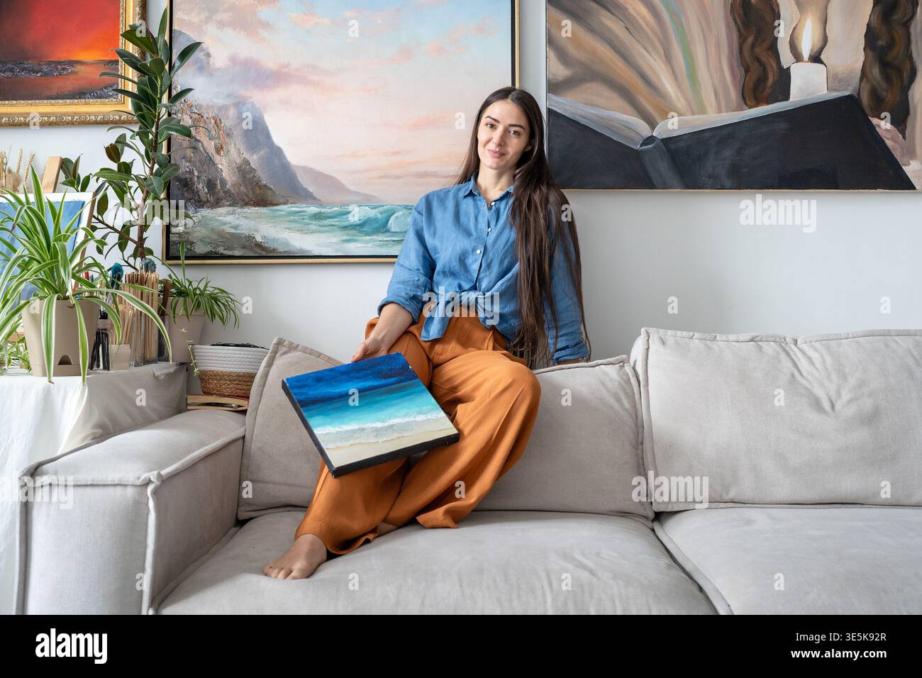 Happy talented woman painter posing at camera with artwork sitting on couch at home loft gallery. Stock Photo