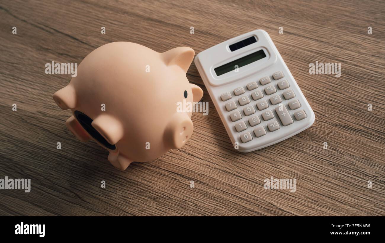 Piggy bank next to a white calculator on wood background for budget planning and accounting metaphors. Stock Photo
