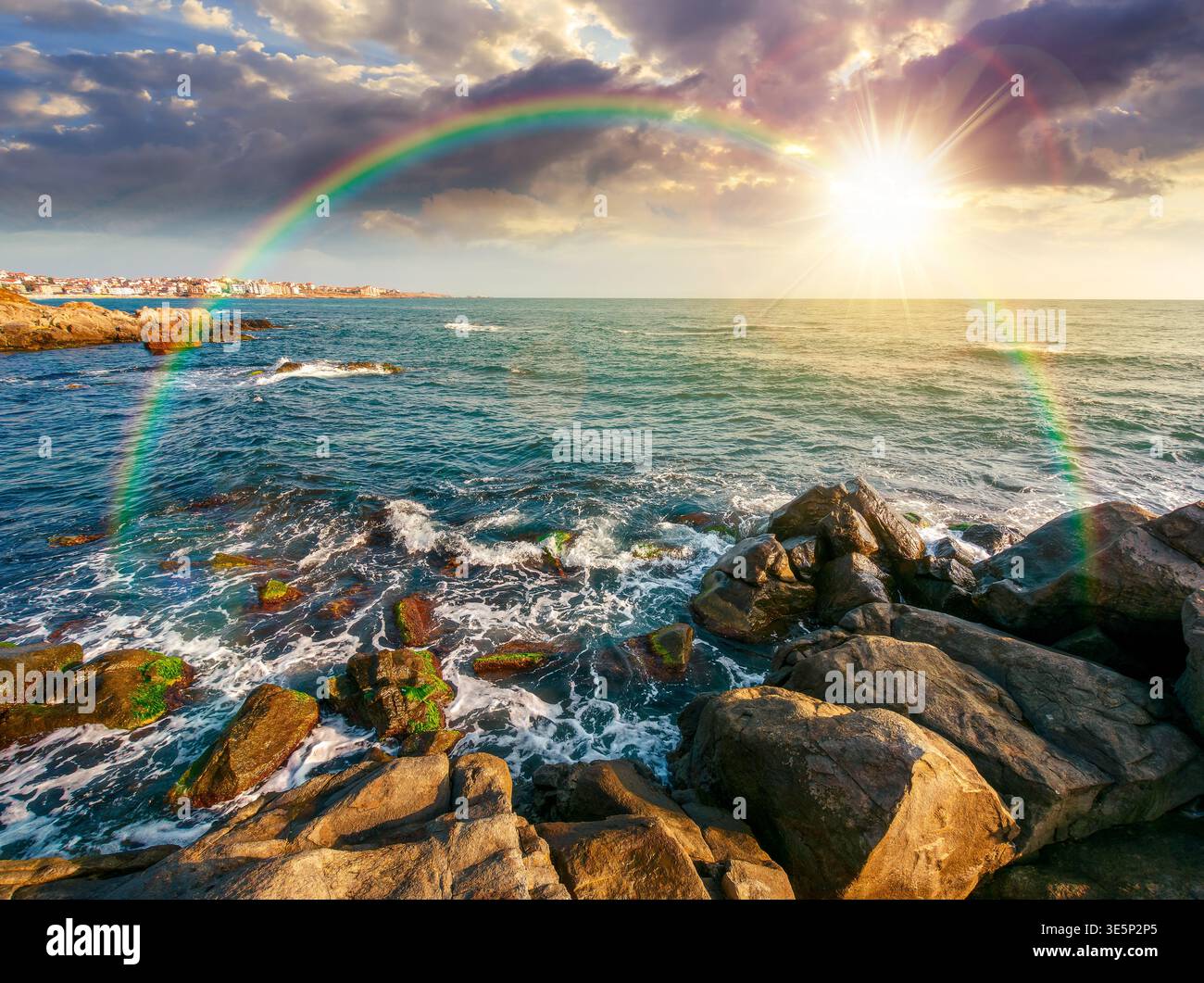 sunset over sea coast with stones and boulders. view of beach with rock in seaweed. dynamic cloudscape on the sky with rainbow above horizon afternoon Stock Photo
