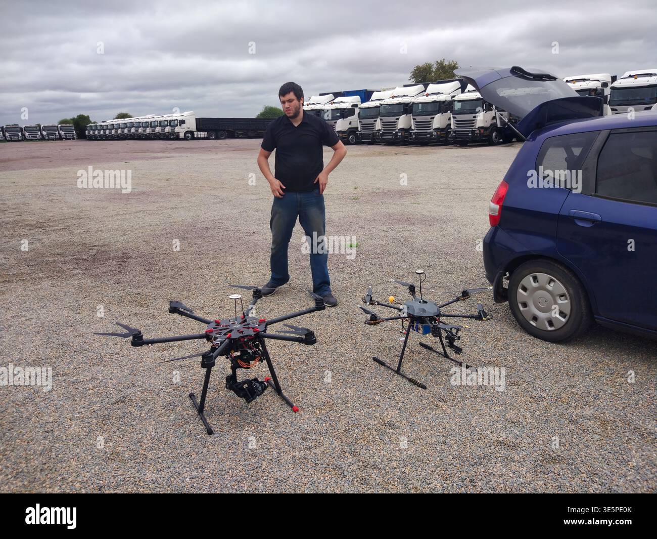 man stands with two professional hexacopter UAVs recently unloaded from the trunk of a blue hatchback in a gravel lot filled with trucks Stock Photo