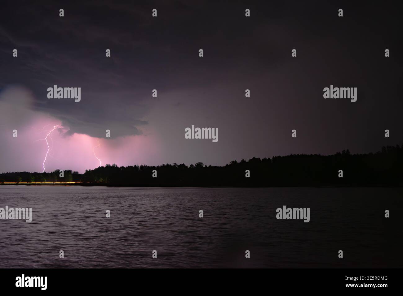 Intense lightning bolts illuminate dark storm clouds over a lake, casting a purple glow across the water and horizon. Stock Photo