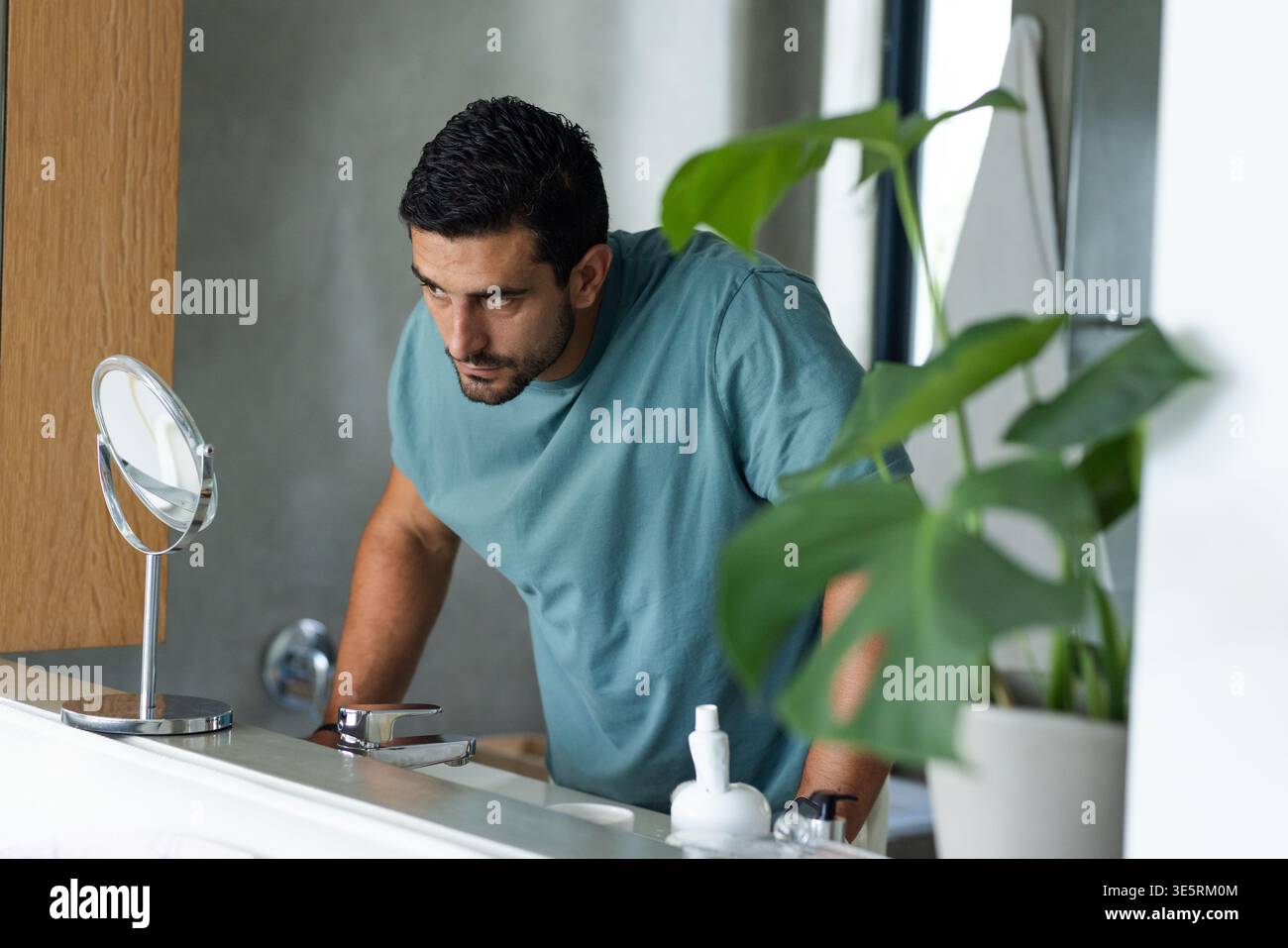 Bearded man wearing teal tee leaning over bathroom sink, looking into mirror while pressing counter Stock Photo