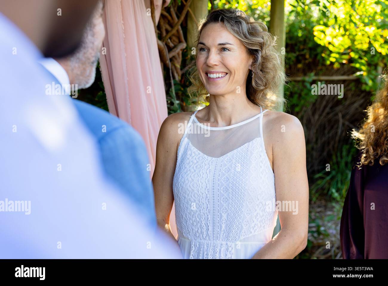 Couple with woman in white sleeveless lace dress smiling under wood arbor with pink drape garden Stock Photo