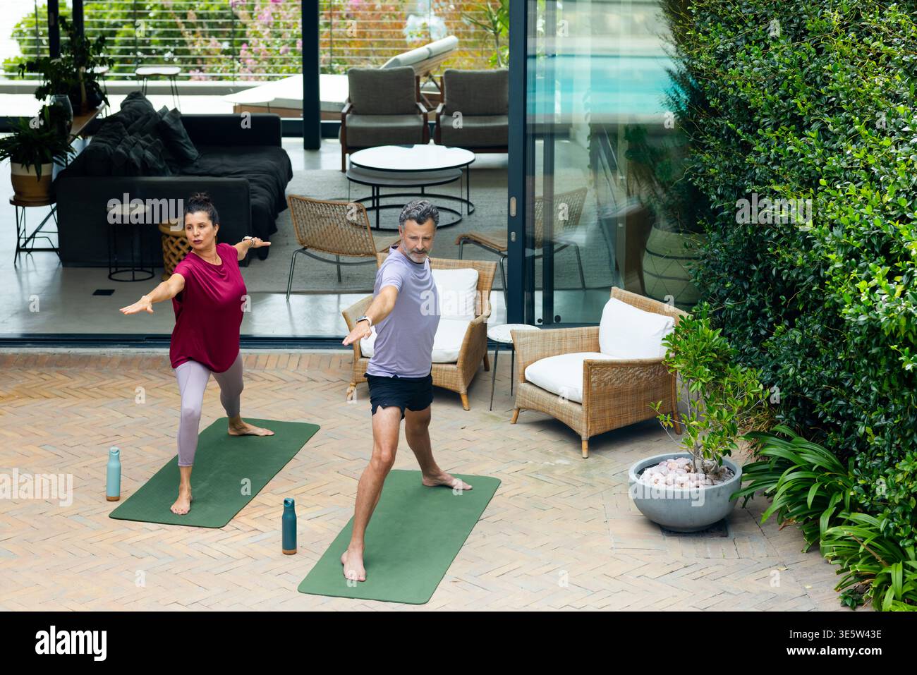 Senior couple doing yoga in activewear on green mats on brick patio near sliding glass doors Stock Photo